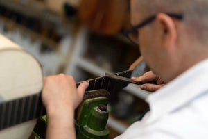 A craftsman carefully shapes the dark fretboard of a guitar using a precision tool.