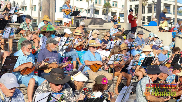 A group of musicians in casual summer attire, many wearing wide-brimmed hats and playing ukuleles.