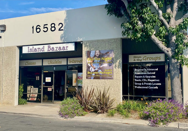 Island bazaar storefront with beige stucco walls, black signage reading ’16582’’ and ’classes ukuleles internet,’ a brown promotional poster for “uke fun night!” featuring a smiling child holding a ukulele