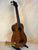 Brown acoustic-electric bass ukulele on a stand, showcasing Kala EM-FS Exotic Macawood Tenor