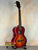 Sunburst-colored acoustic guitar on black stand, featuring Kala KA-FMCB-T gloss flame maple Cherry Burst