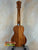 Wooden ukulele with decorative headstock on stand, showcasing KoAloha KCM-00 Concert design