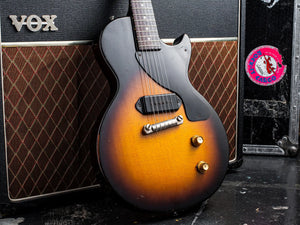 Sunburst-finished electric guitar with a black pickguard and gold hardware, leaning against a vintage vox amplifier.