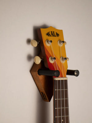 A vibrant yellow and orange gradient kala ukulele headstock with cream-colored tuning pegs mounted on a wooden wall stand.